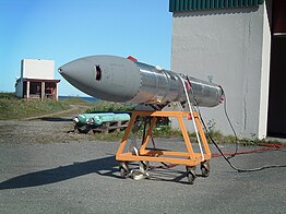 An elongated cyllindric object, ending with a cone, placed on a small, openwork trolley, in front of a hangar, on a sunny day.