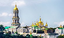 Panoramablick auf das Kloster mit den Häusern im Vordergrund, alles mit weißen Fassaden und goldenen Kirchtürmen nach orthodoxer Architektur. Auf der linken Seite ist ein großer Kirchturm mit drei Etagen zu erkennen, in der Mitte befinden sich weiter im Hintergrund mehrere Kirchtürme.