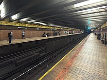 People waiting on a dimly lighted, nearly empty brick-tile platform at the 21st Street–Queensbridge station