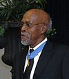 A gray-haired black man standing at a microphone, wearing a medal on a blue ribbon around his neck.