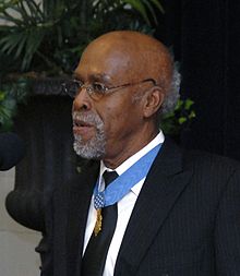 Clarence Sasser, a gray-haired black man standing at a microphone, wearing a medal on a blue ribbon around his neck.