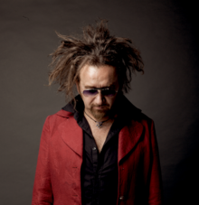 Portrait d'un homme d'une soixantaine d'années portant les cheveux longs en dreadlocks et un bouc, avec une veste rouge, une chemise noire et des lunettes de soleil.