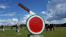 Several scrap metal sculptures in a field of grass, including a watermelon being cut by a knife and a life-size elephant.