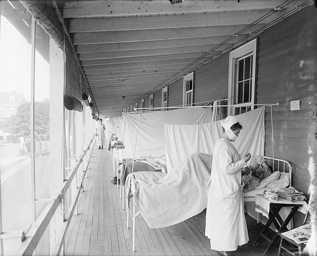 mehrere Krankenbetten, von an der Spanischen Grippe Erkrankten, welche nebeneinander auf einem überdachten Balkon stehen, getrennt durch weiße Laken. Eine Krankenschwester mit Mundschutz versorgt einen Patienten
