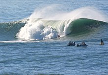 Photo of taller-than-human-sized wave breaking with several watching surfers in foreground
