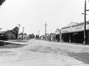 black and white photo of a street with buildings and power lines on either side and a rail line crossing lower right to mid left; station building on the left, mid frame