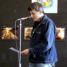 a man in a denim jacket reads off pages into a microphone on a stand