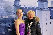 Viveca Lindfors trägt lilafarbenes ärmelloses Eiskunstlaufkleid. Ihre Trainerin, die den Arm um ihre Schultern gelegt hat, trägt eine dunkle Winterjacke. Hinter ihnen ein Stadtpanorama.