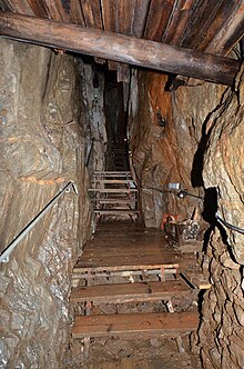 A narrow passageway inside the mine, with rocks to the left and right, wooden stairs at the bottom, and wooden rafters above