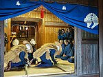 Two men in straw coats bowing on a tatami floor inside a house.