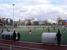 Des footballeurs et un arbitre courent sur un terrain au cours d'un match, devant un petit nombre de personnes se tenant derrière une main courante.