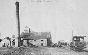 Une locomotive vue de derrière passant sur une voie ferrée entourée de bâtiments industriels.