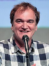 A smiling Quentin Tarantino dressed in a casual shirt speaking into a microphone