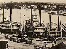 A black-and-white image showing a large river with four steamships docked by the shore. Numerous wooden buildings appear on both sides of the river.
