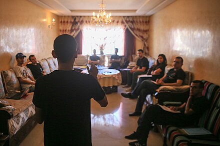 Photo is taken from behind Reda, there are 8 participants listening sat on couches around the outside of a rectangle room with a window at the far end