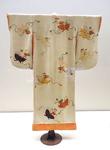 Vêtement de forme kimono, en crêpe écru, portant des broderies dans des tons orange, jaune et marron, représentant des papillons et des chrysanthèmes.