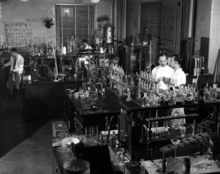 Photograph of a room with multiple scientists in white lab coats standing among tables covered in glassware