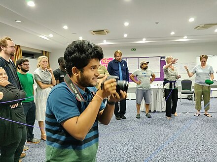 Participants in a circle with yarn connecting each other. A photographer is centre frame about lifting a camera to his face.