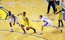 A basketball player in a maize colored uniform dribbles between defenders in white uniforms.