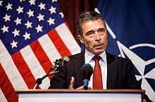 An older man in a suit speaking at a podium in front of two flags.