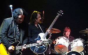 Prominent in the foreground, a guitarist concentrates on his playing, while the drummer, a little behind him to his left, toils away.