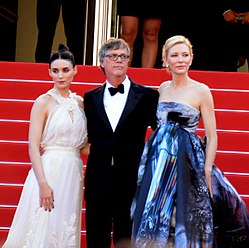A man wearing a black tuxedo, white shirt, black bow tie, and eyeglasses is standing with his arms around the backs of two women. Behind them are red-carpeted steps. The woman on his right, a brunette, is wearing an haute couture white halter gown with ruffles and embroidery on the skirt; while the woman on his left, a blonde, is wearing a strapless ballgown with prints and shades of blue, light gray, black, and some hints of red.
