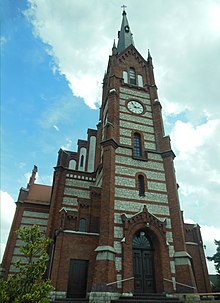 Ceglana wieża zdobiona białym kamieniem oraz główne wejście do kościoła, widoczny zegar i iglica wieży, portal i drzwi boczne