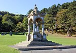 The monument; a bust on a pedestal covered by a free-standing canopy, behind which is a lawn with Gorsedd stones