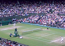 Two men dressed in white, seen from a distance, play on a grasscourt in a crowded stadium, at dusk