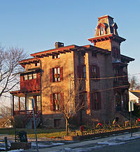 A brick house with a peaked tower and red wooden trim, seen from far to its right and illuminated by late-afternoon sun from the left