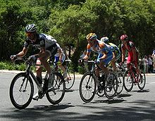 A group of five cyclists on the road. Three, in the foreground, are crouched over their bicycles, while the other two are riding out of the saddle. Spectators are visible on the roadside.