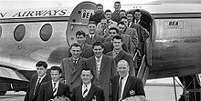 A black-and-white photograph of several people  in suits and overcoats on the steps of an aircraft.