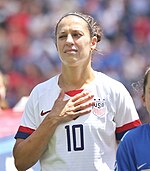 Photographie de l'attaquante américaine Carli Lloyd portant le maillot de l'équipe des États-Unis et posant sa main sur son cœur durant la diffusion de l'hymne américain.