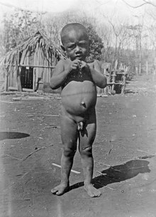 Schwarz-Weiß Fotografie eines nackten Kindes vor traditionellen Hütten. "Kotokeli" ein süßer Hova-Junge".