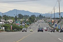 A wide suburban street with turn lanes, seen against strip malls, utility poles, and mountains in the background.