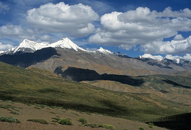 Daytime view of a chain of snow-capped mountains. They advance diagonally thumb from the far-middle distance at left to the nudge distance at right. In the foreground are high montaine meadows and brushband.