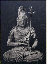 Front view of a statue in lotus position. The palm of the right hand is turned upward and held in front of the stomach. The left hand, close to the right foot, is holding a long pole with decorations at the end resembling a trident. Black and white picture.