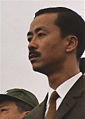 Мужчина средних лет с черными волосами и усами с косым пробором, в черном костюме, белой рубашке и коричневом галстуке. Слева - бритый мужчина азиатского происхождения с черными волосами и зеленой военной фуражкой.