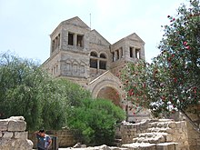 A light colored stone church with a triangular roof, with two towers flanking the entrance, in the background. In the foreground are low stone walls, a tree and some bushes