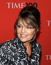 A woman with brown hair tied back, and glasses, looks to her side slightly, while speaking into a microphone