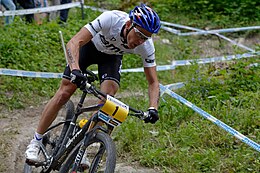 Homme descendant une pente à VTT, portant un casque bleu, un maillot blanc et un cuissard noir.