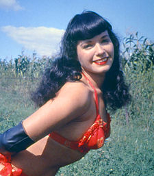 Černovlasá Bettie Page se s úsměvem nahýbá zleva a zblízka do záběru fotografa, na sobě má červené dvoudílné plavky a v pozadí je pole se vzrostlou kukuřicí
