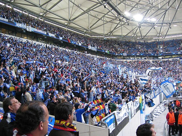 A pitch level perspective of the corner section of a stadium filled with people holding blue scarfs and waving blue-and-white flags.
