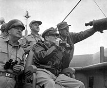 MacArthur sedí, nosí klobouk svého polního maršála a bombardovací bundu a drží dalekohled. Čtyři další muži nesoucí dalekohled stojí za ním.