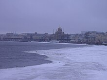 Photo aux tons gris-mauve, comme brumeux ; au fond, des bâtiments ; devant, une grande étendue grise et blanche