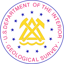 The Seal of the United States Geological Survey. The central element is a crossed pick and hammer, a traditional symbol of mining, over a stylized symbol of a mountain and water.