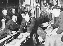 A group of men in woollen jerseys, several smoking pipes, are watching repair work on a sledge. They are in a confined area, with equipment and spare clothing adorning the walls