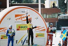 Trois fondeurs de face sur un podium, celui du centre brandissant de son bras droit un trophée en forme de globe de cristal.