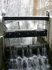 Weir in place of the upper doors of the lock. This weir has a levelling mechanism attached, rather like a tilting weir. We see the dark frames of the weir, wet on a winter's day, with the water cascading over toward us in a smooth cataract, no turbulence or disturbance. The surface of the mill pond beyond is placid. The air is misty and the surrounding trees are white with hoar-frost, which also rimes the edges of the frame of the weir, melted only where the water has splashed.