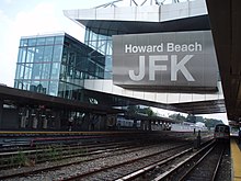 Blick von einem Bahnsteig auf eine brückenartiger Überbauung über den Gleisen, beschriftet mit dem übergroßen Schriftzug „Howard Beach JFK“